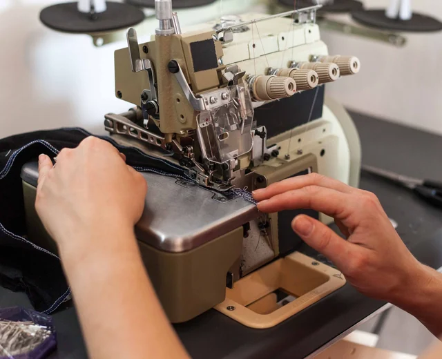 Hands guiding a dark knit panel through an overlock machine on the sewing line