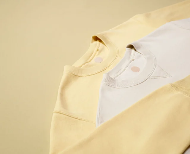 Cream and yellow sweatshirts laid flat, showing rib necklines and blank printed neck labels