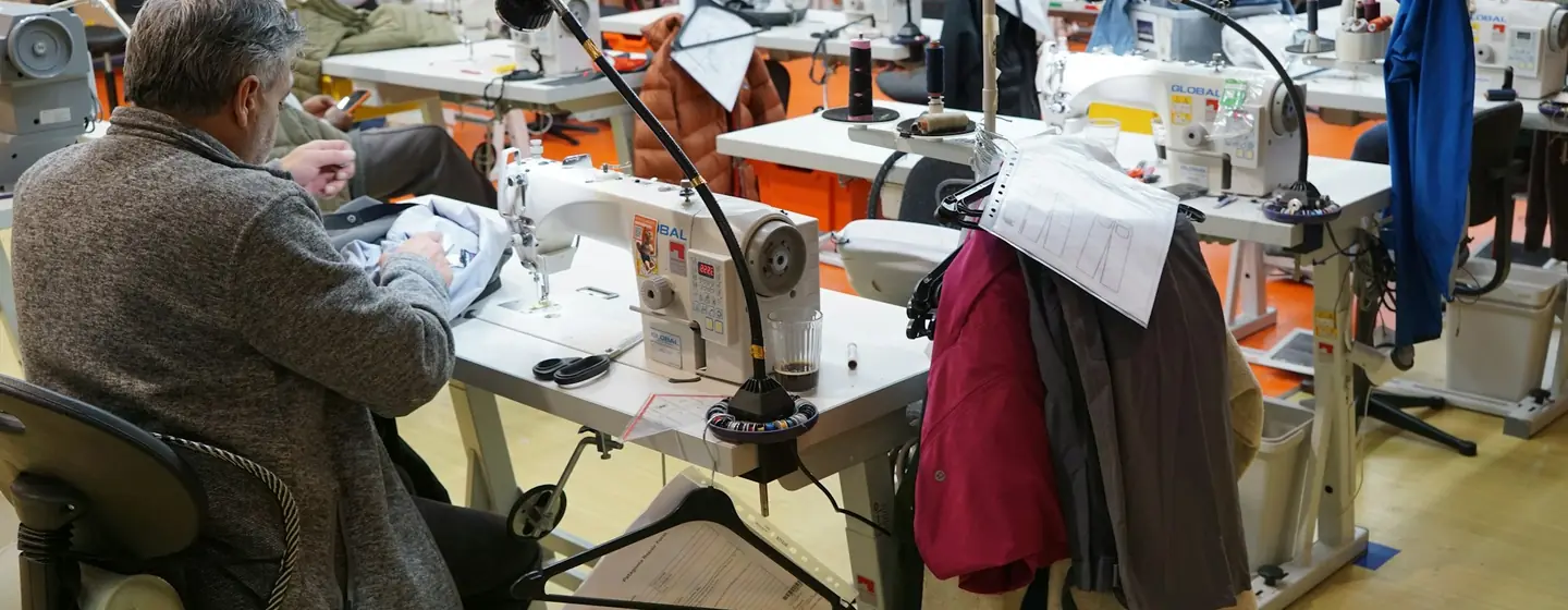 Sewing operator working on jackets in a busy garment workshop with rows of industrial machines